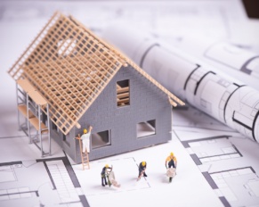 Arkusz papieru z planami budynku, na mim stoi model domu w budowie, obok ustawione małe figurki budowlańców