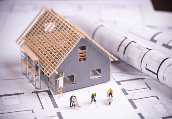 Arkusz papieru z planami budynku, na mim stoi model domu w budowie, obok ustawione małe figurki budowlańców